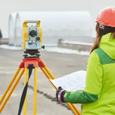 Geodeta w kasku i kamizelce, z mapą w ręku, pracuje przy tachimetrze. Tak, geodeta może wejść na posesję w celu wykonania pomiarów.
