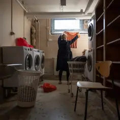 Kobieta wiesza pranie w suterenie. W pralni stoją pralki, kosze na pranie i puste półki.