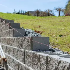 Budowa muru oporowego z bloczków betonowych na zboczu. Widać drabinę i różową nić traserską.