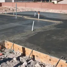 Świeżo wylany beton na płycie fundamentowej, widoczne rury i zbrojenie. Widać przygotowania do izolacji pionowej fundamentów.