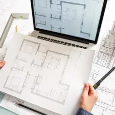 Architekt analizuje plany mieszkania, które mogą być jego pierwsze klucze do sukcesu. Na stole leżą projekty, laptop i makieta domu.