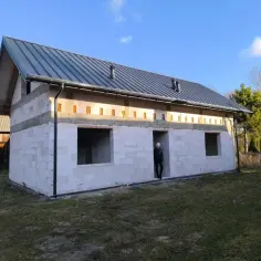 Budowa domu na zgłoszenie w stanie surowym, z betonowych pustaków i metalowym dachem. W drzwiach stoi osoba.
