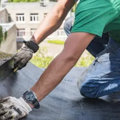 Dekarz w rękawicach i zielonej koszulce pracuje na dachu, układając papę.
