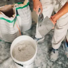 Ręce tynkarza mieszają masę, obok worki z materiałem. Sprawdź aktualne tynki cementowo wapienne cena.