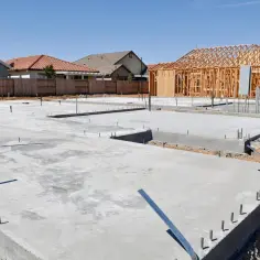 Fundamenty domów w budowie, widać chudy beton i zbrojenia. W tle drewniana konstrukcja domu.