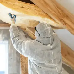 Pracownik w kombinezonie izoluje poddasze nieużytkowe wełną mineralną.