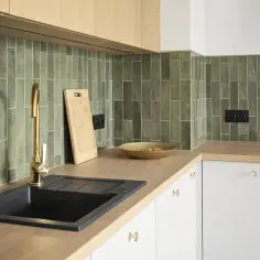 Drewniane blaty kuchenne w nowoczesnej kuchni z zieloną, ceglaną mozaiką. Widoczny złoty kran, czarny zlew i deska do krojenia.