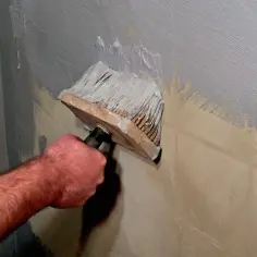 Ręka z pędzlem nakłada białą farbę na szarą ścianę. Czy można malować farbą ceramiczną na lateksową?