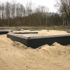 Budowa domu w piasku, widoczne ocieplenie fundamentów czarną folią. W tle las.