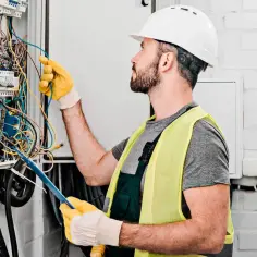 Elektryk w kasku i kamizelce odblaskowej pracuje przy skrzynce z przewodami. Sprawdza instalację, szukając informacji o montaż gniazdka cena.