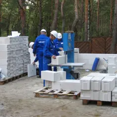 Budowa domu z bloczków betonowych. Na placu budowy pracownicy przygotowują materiał, a stosy bloczków sugerują, ile bloczków betonowych na m2 potrzeba do wzniesienia ścian.