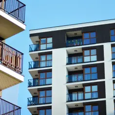 Nowoczesne budynki z loggią i balkonem, z jasnymi fasadami i ciemnymi akcentami, na tle błękitnego nieba.