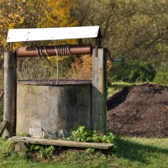Stara studnia kręgowa z drewnianą konstrukcją i korbą, otoczona jesienną zielenią i stertą ziemi.