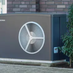 Nowoczesna pompa ciepła, szary panel zewnętrzny z okrągłym wiatrakiem, otoczona zielenią. Sprawdź, jaki pompa ciepła koszt.