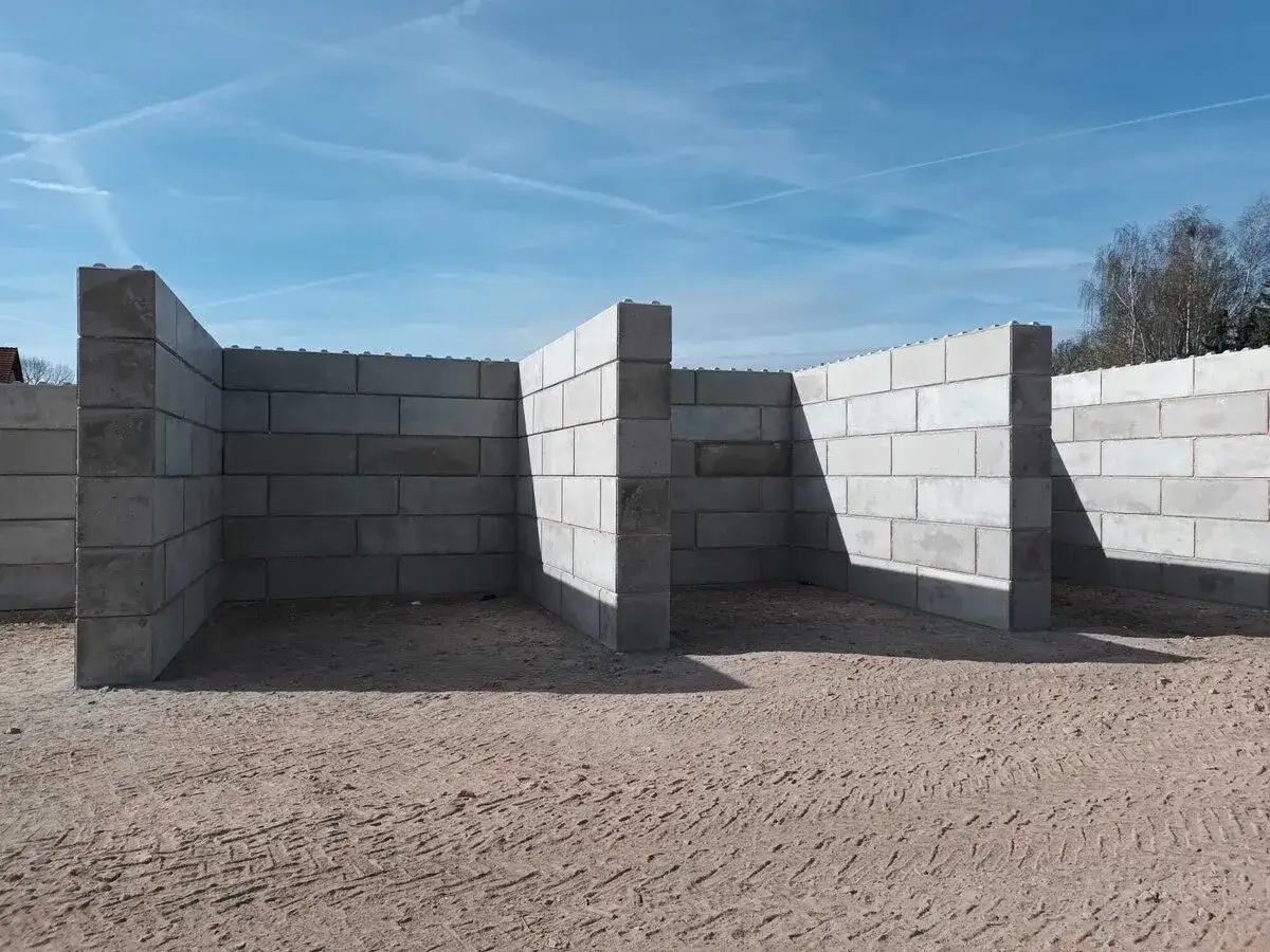 Budowa muru oporowego z bloczków betonowych. Konstrukcja tworzy zarys pomieszczeń na piaszczystym placu budowy.