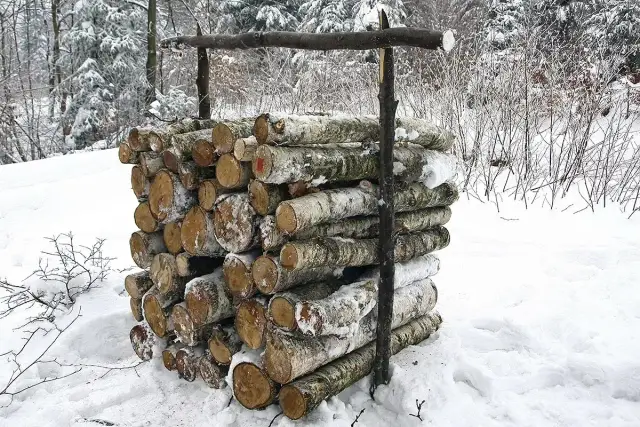Zimowy stos drewna, prawdopodobnie metr sześcienny, przygotowany do palenia. Śnieg pokrywa pnie i otoczenie.